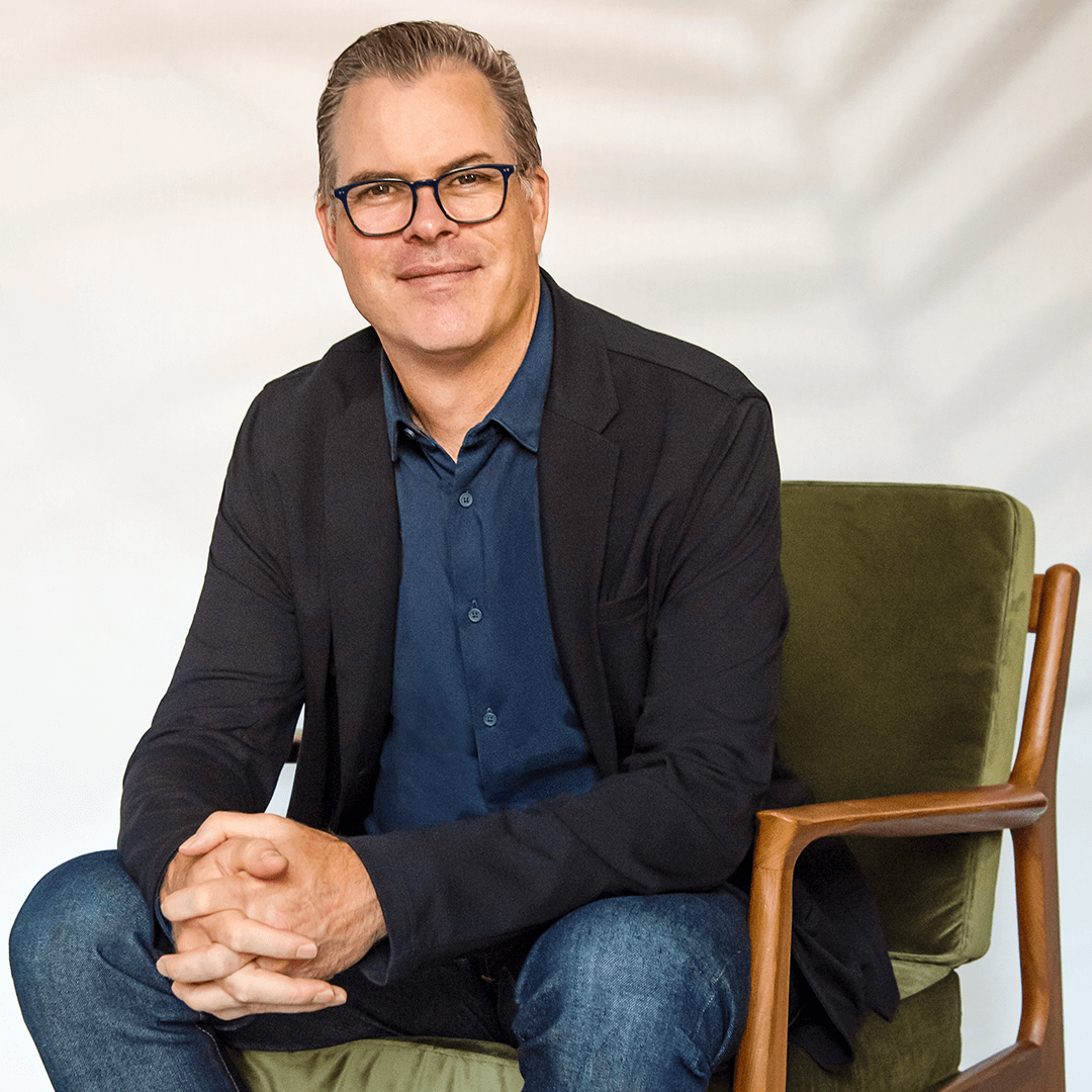 Dave Nourse smiling and sitting in a vintage-style wooden armchair with green cushions with his hands clasped together.