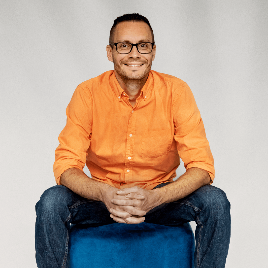 François Mouchet sitting in a square pouf with hands clasped together.