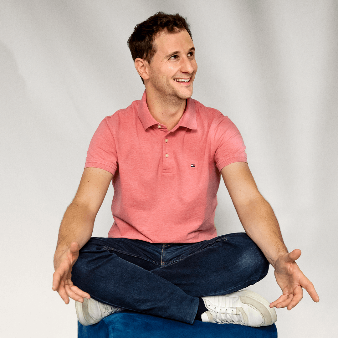 Fréderic Vassal sitting cross-legged on a blue cube, smiling and looking to the side.