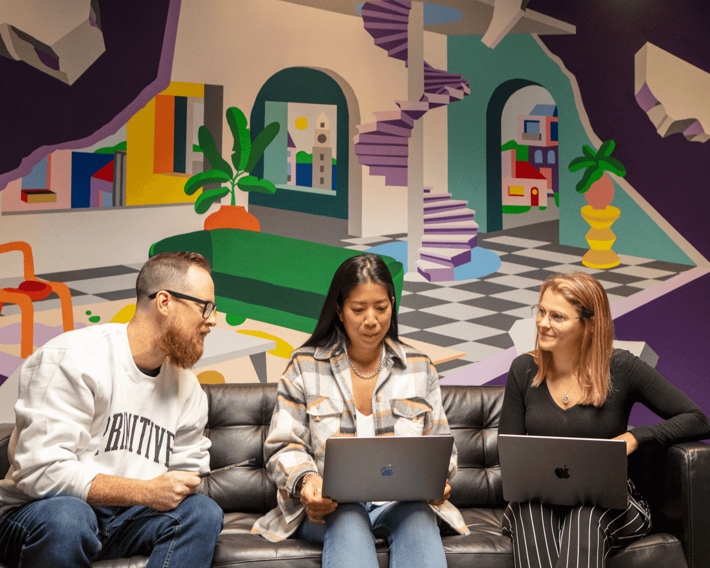 m&h employees collaborating on laptops, seated in front of a colourful wall mural in the office