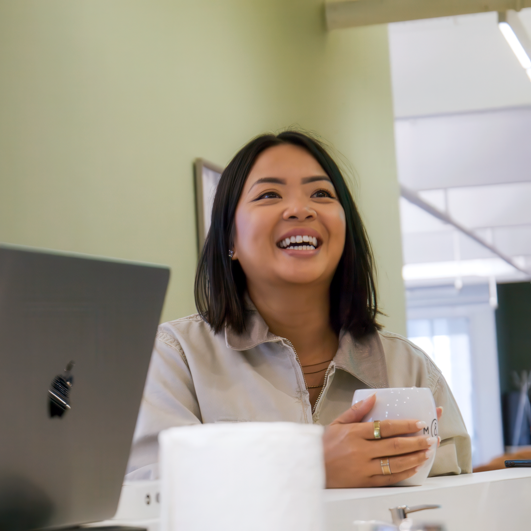 Employée m&h souriante, tasse à la main dans un bureau lumineux.
