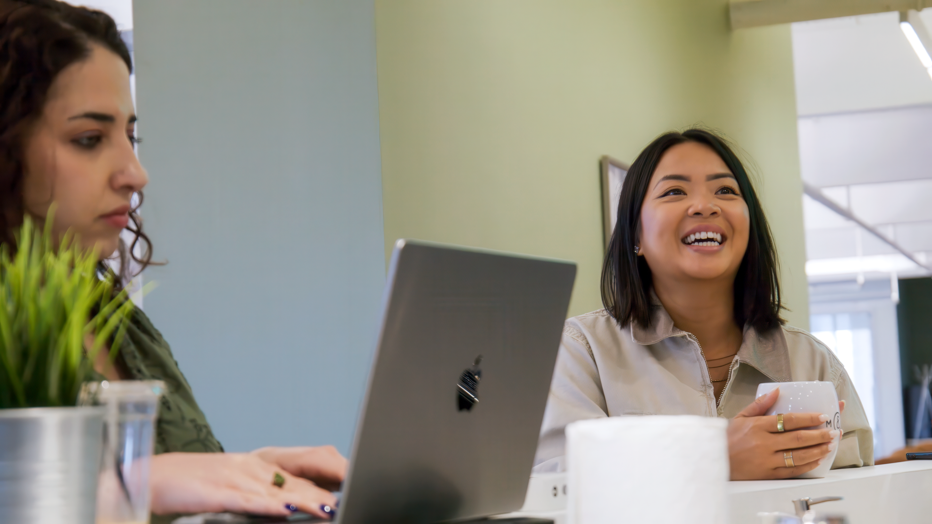 Two women in a relaxed office setting, one smiling with a cup in hand and the other focused on her laptop.